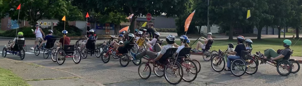 20+ people on various adaptive handcycles, tricycles, tandems, side-by-side bikes, or therapy trikes (many with flags for visibility). 20+ people on various adaptive handcycles, tricycles, tandems, side-by-side bikes, or therapy trikes (many with flags for visibility).